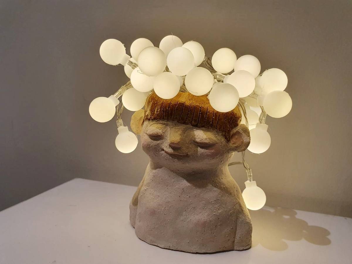 周宛螢的陶藝作品「小聰明」，在表情恬靜安逸的陶偶頭頂裝上許多暖色燈泡，像一顆顆在童年發光的自信與期待，是內心仍保有孩子般對世界最純真的盼望。